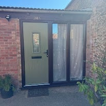 Exterior view of a green entry door on a brick building with glass panels and potted plants on the step