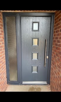 Modern gray front door with four square glass panes mounted on a brick wall with a side panel window