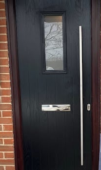 Black front door with rectangular window and silver handle mounted on red brick wall