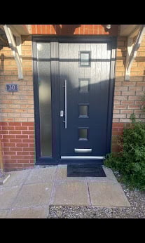 Modern black front door with chrome handle on a brick house entrance with paved walkway and green shrub