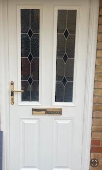 White front door with two glass panes featuring geometric lattice patterns, brass handle, and nameplate